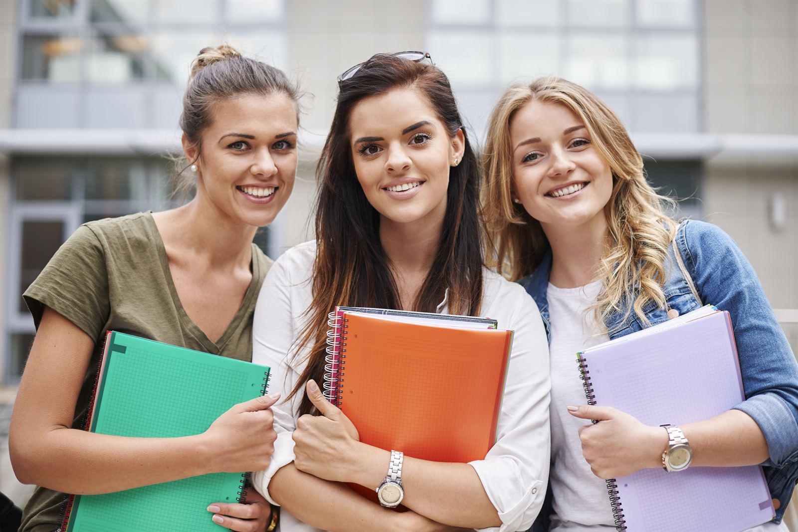Three smiling students holding notebooks outside a school building.