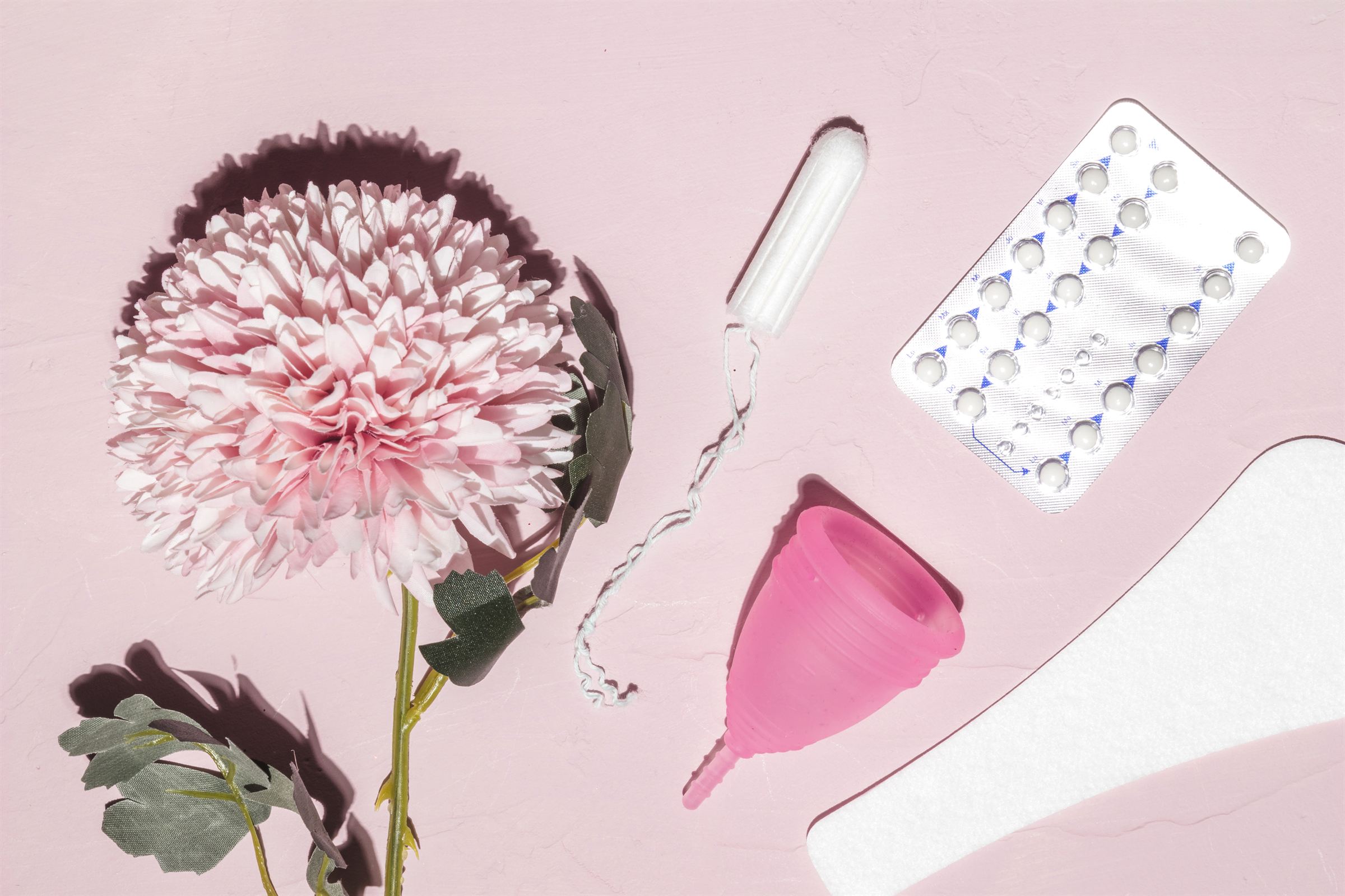 A pink flower, a tampon, a menstrual cup, a pad and a blister pack of pills laid out on a soft pink background.